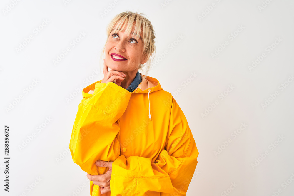 Middle age woman wearing yellow rain coat standing over isolated white background with hand on chin thinking about question, pensive expression. Smiling with thoughtful face. Doubt concept. Middle age woman wearing yellow rain coat standing over isolated white background with hand on chin thinking about question, pensive expression. Smiling with thoughtful face. Doubt concept.