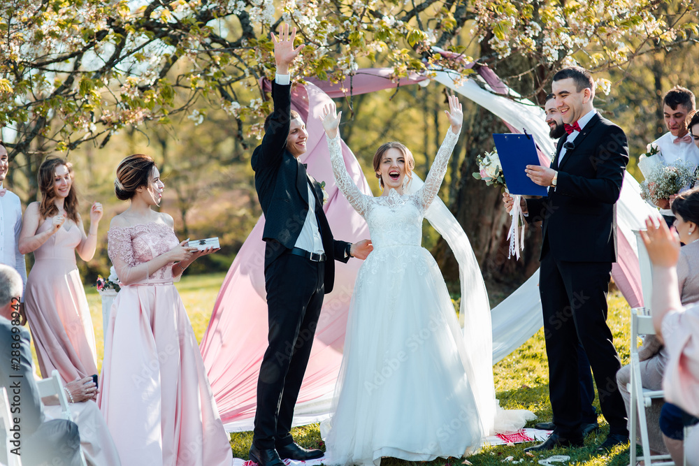 Bride and groom after wedding ceremony. The master of ceremonies reads from a sheet beautiful words to newlyweds who are standing in green sunny park. Stylish happy smiling newlyweds on the outdoor
