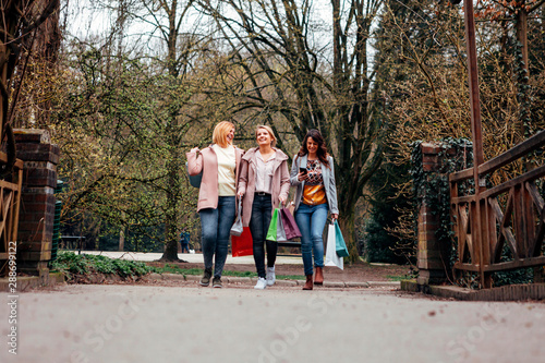 Fototapeta Naklejka Na Ścianę i Meble -  three happy and smiling women walk together carrying shopping bags and using a mobile phone