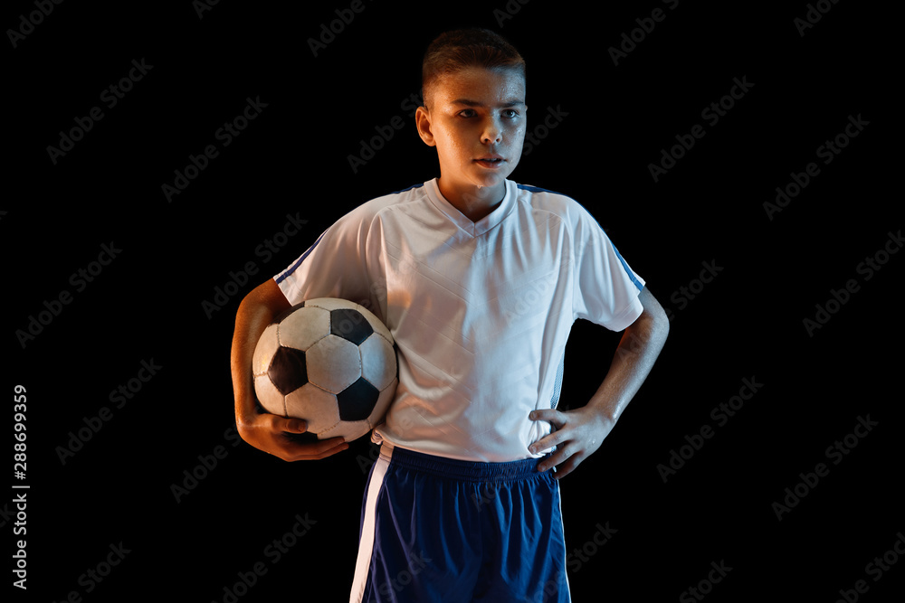 Young boy as a soccer or football player in sportwear making a feint or a kick with the ball for a goal on dark background. Fit playing boy in action, movement, motion at game. Confident and strong. Young boy as a soccer or football player in sportwear making a feint or a kick with the ball for a goal on dark background. Fit playing boy in action, movement, motion at game. Confident and strong.
