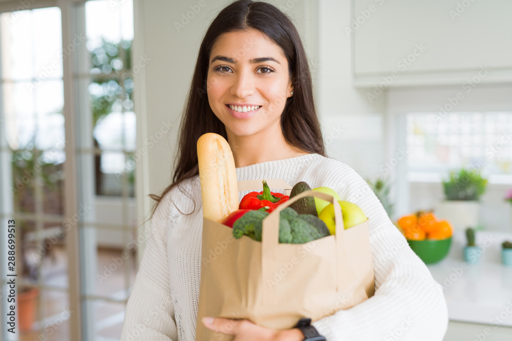 Beautiful young woman smiling holding a paper bag full of groceries at the kitchen Beautiful young woman smiling holding a paper bag full of groceries at the kitchen