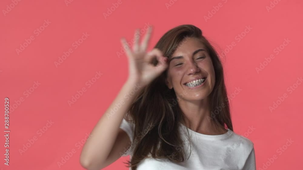 Attractive cheerful adult brunette woman with braces in white t-shirt, spend excellent time, having fun, joyfully smiling show okay sign, laughing from happiness and joy, standing red background Attractive cheerful adult brunette woman with braces in white t-shirt, spend excellent time, having fun, joyfully smiling show okay sign, laughing from happiness and joy, standing red background