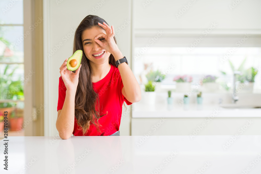 Young woman eating healthy avocado with happy face smiling doing ok sign with hand on eye looking through fingers Young woman eating healthy avocado with happy face smiling doing ok sign with hand on eye looking through fingers