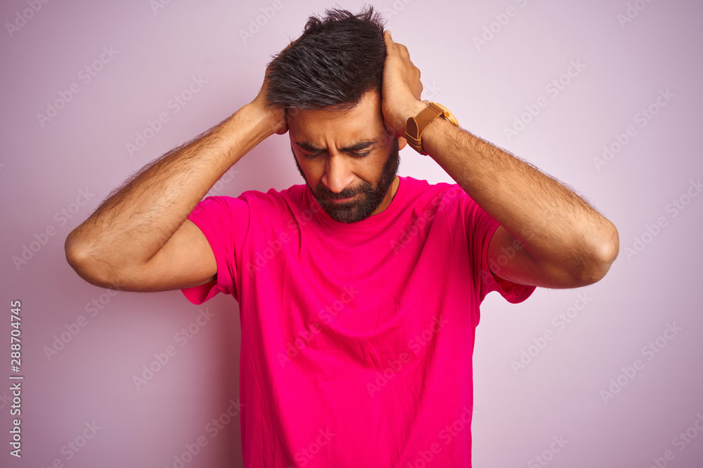 Young indian man wearing t-shirt standing over isolated pink background suffering from headache desperate and stressed because pain and migraine. Hands on head. Young indian man wearing t-shirt standing over isolated pink background suffering from headache desperate and stressed because pain and migraine. Hands on head.