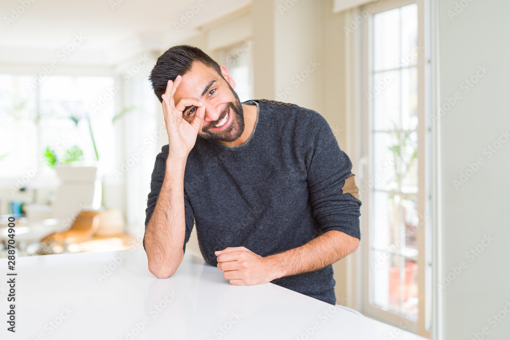 Handsome hispanic man wearing casual sweater at home doing ok gesture with hand smiling, eye looking through fingers with happy face.