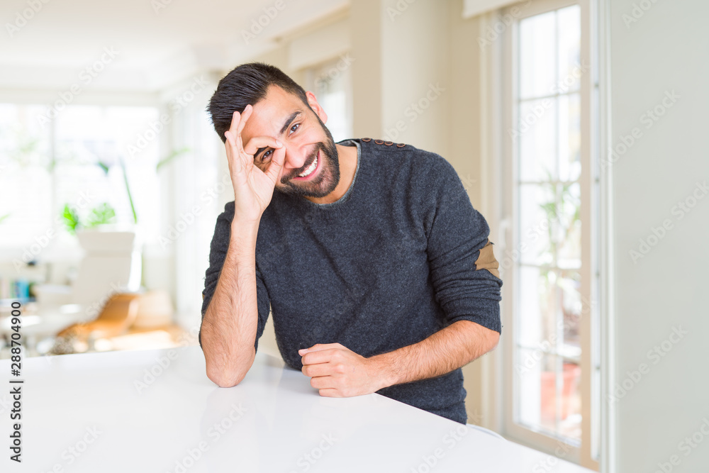 Handsome hispanic man wearing casual sweater at home doing ok gesture with hand smiling, eye looking through fingers with happy face. Handsome hispanic man wearing casual sweater at home doing ok gesture with hand smiling, eye looking through fingers with happy face.