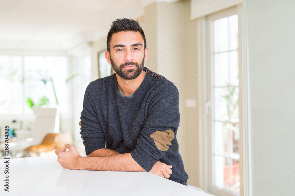 Handsome hispanic man wearing casual sweater at home with serious expression on face. Simple and natural looking at the camera.