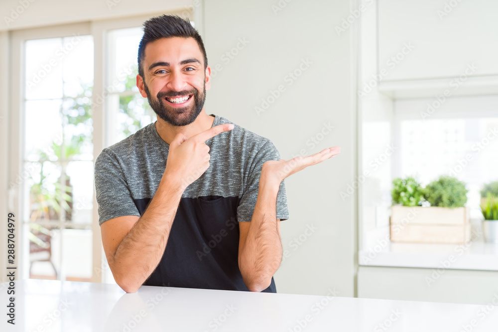Fototapeta premium Handsome hispanic man wearing casual t-shirt at home amazed and smiling to the camera while presenting with hand and pointing with finger.