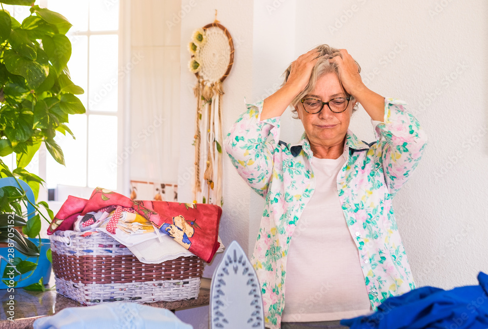 Obraz premium A senior woman with grey hair sad and disperate because she has too much clothes to iron. Corner of the domestic room with plants and white wall. One people only