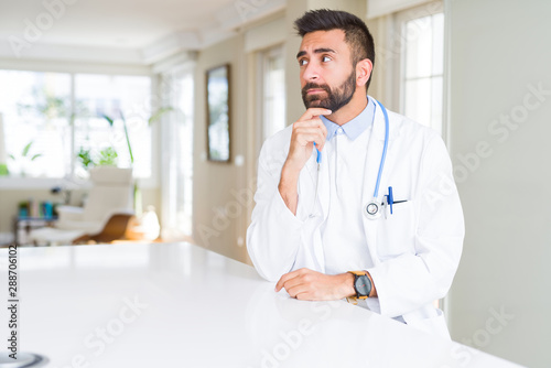 Handsome hispanic doctor man wearing stethoscope at the clinic with hand on chin thinking about question, pensive expression. Smiling with thoughtful face. Doubt concept.