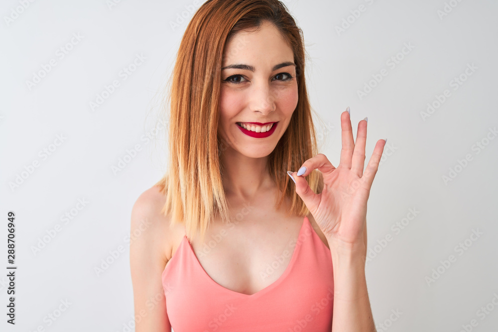 Beautiful redhead woman wearing casual pink t-shirt standing over isolated white background doing ok sign with fingers, excellent symbol Beautiful redhead woman wearing casual pink t-shirt standing over isolated white background doing ok sign with fingers, excellent symbol