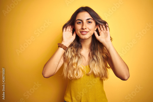 Young beautiful woman wearing t-shirt over yellow isolated background Trying to hear both hands on ear gesture, curious for gossip. Hearing problem, deaf
