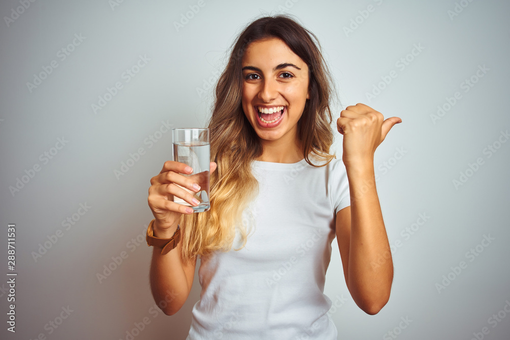 Young beautiful woman drinking a glass of water over white isolated background pointing and showing with thumb up to the side with happy face smiling Young beautiful woman drinking a glass of water over white isolated background pointing and showing with thumb up to the side with happy face smiling