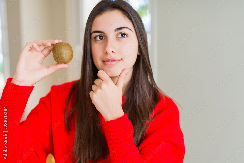 Beautiful young woman eating fresh green kiwi serious face thinking about question, very confused idea Beautiful young woman eating fresh green kiwi serious face thinking about question, very confused idea
