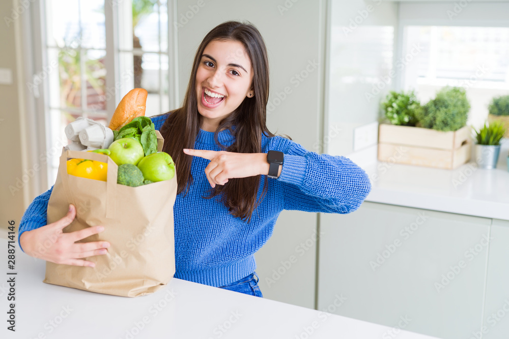 Beautiful young woman holding paper bag full of healthy groceries very happy pointing with hand and finger Beautiful young woman holding paper bag full of healthy groceries very happy pointing with hand and finger