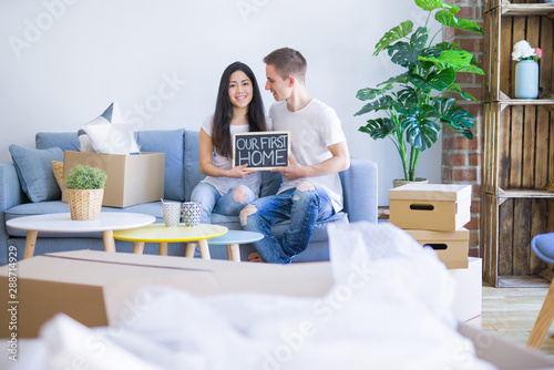 Young beautiful couple sitting on the sofa holding blackboard with message at new home around cardboard boxes