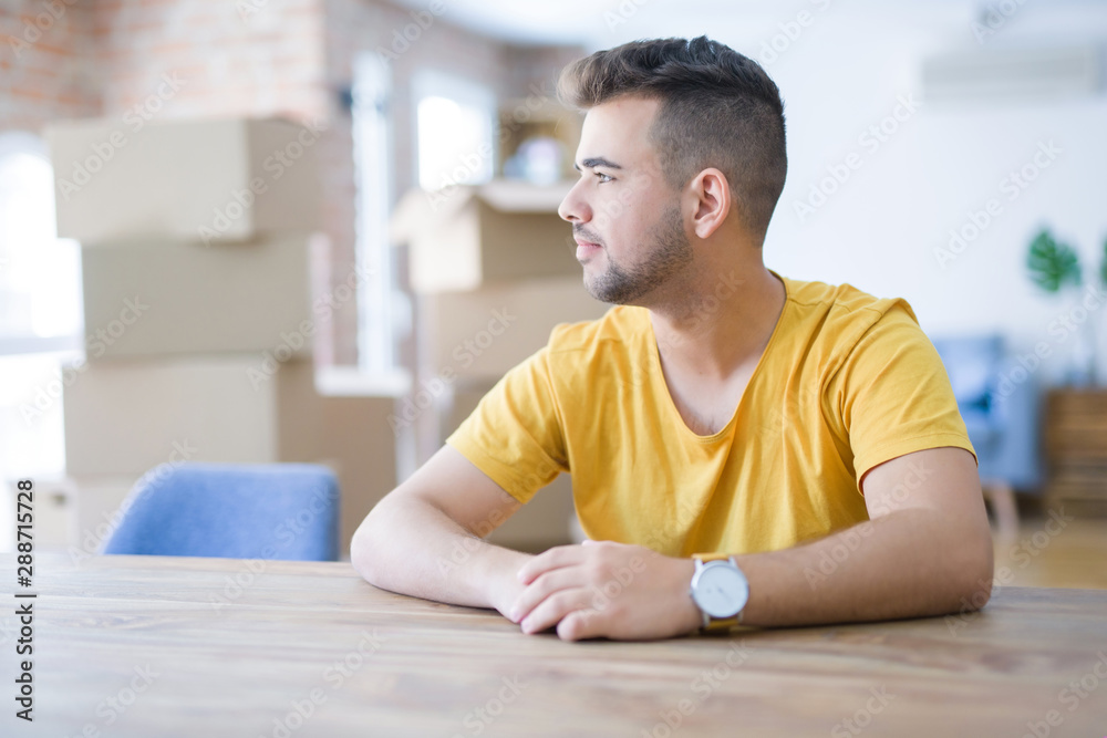 Young man sitting on the table with cardboard boxes behind him moving to new home looking to side, relax profile pose with natural face with confident smile. Young man sitting on the table with cardboard boxes behind him moving to new home looking to side, relax profile pose with natural face with confident smile.