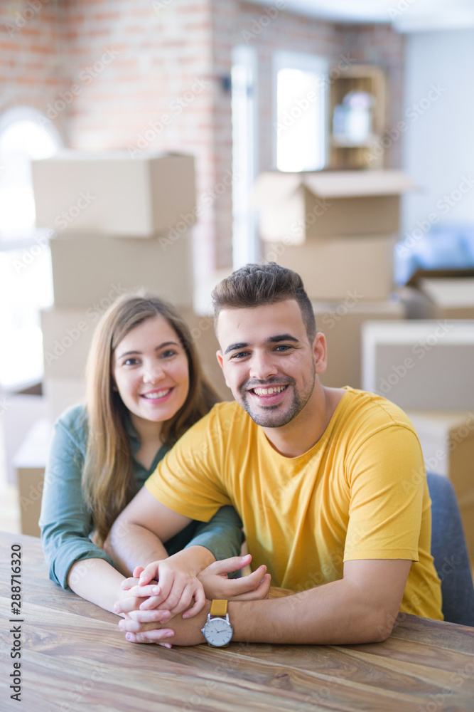 Young beautiful couple sitting on the table at home, hugging in love very happy for moving to new home with cardboard boxes behind them Young beautiful couple sitting on the table at home, hugging in love very happy for moving to new home with cardboard boxes behind them