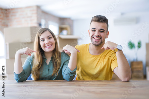 Young couple sitting on the table movinto to new home with carboard boxes behind them looking confident with smile on face, pointing oneself with fingers proud and happy.