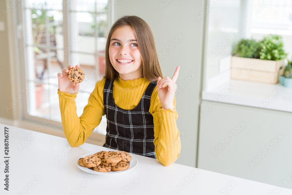 Beautiful young girl kid eating chocolate chips cookies very happy pointing with hand and finger to the side Beautiful young girl kid eating chocolate chips cookies very happy pointing with hand and finger to the side