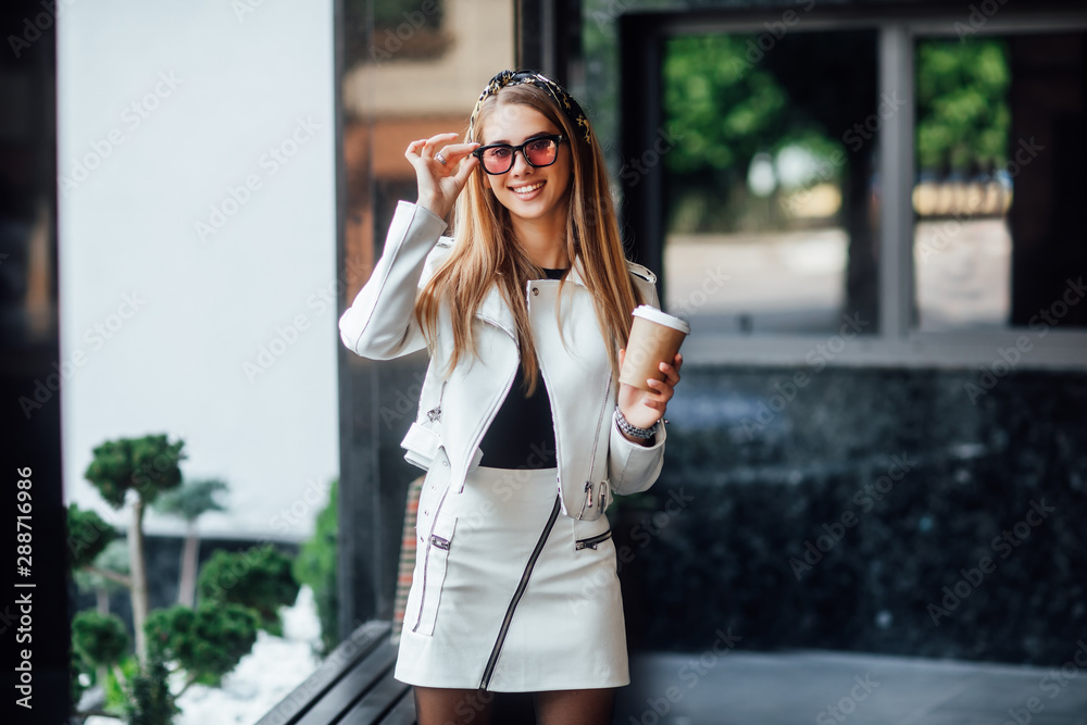Portrait of young blonde model walking in the city, girl in stylish outfit drinking coffe and enjoying.