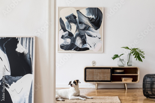 Design scandi home interior of living room with wooden commode, gray sofa, black rattan pouf, plant and elegant accessories. Stylish home decor. Mock up abstract paintings. Dog is lying on the carpet