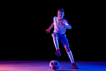 Young boy as a soccer or football player in sportwear making a feint or a kick with the ball for a goal on dark studio background. Fit playing boy in action, movement, motion at game. Purple neon