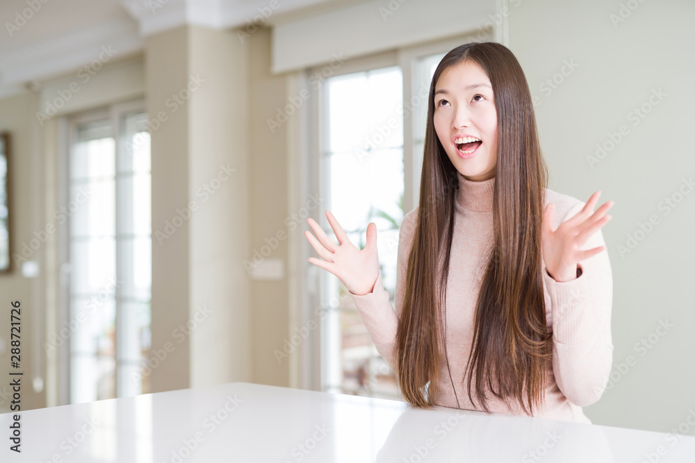Beautiful Asian woman wearing casual sweater on white table crazy and mad shouting and yelling with aggressive expression and arms raised. Frustration concept. Beautiful Asian woman wearing casual sweater on white table crazy and mad shouting and yelling with aggressive expression and arms raised. Frustration concept.