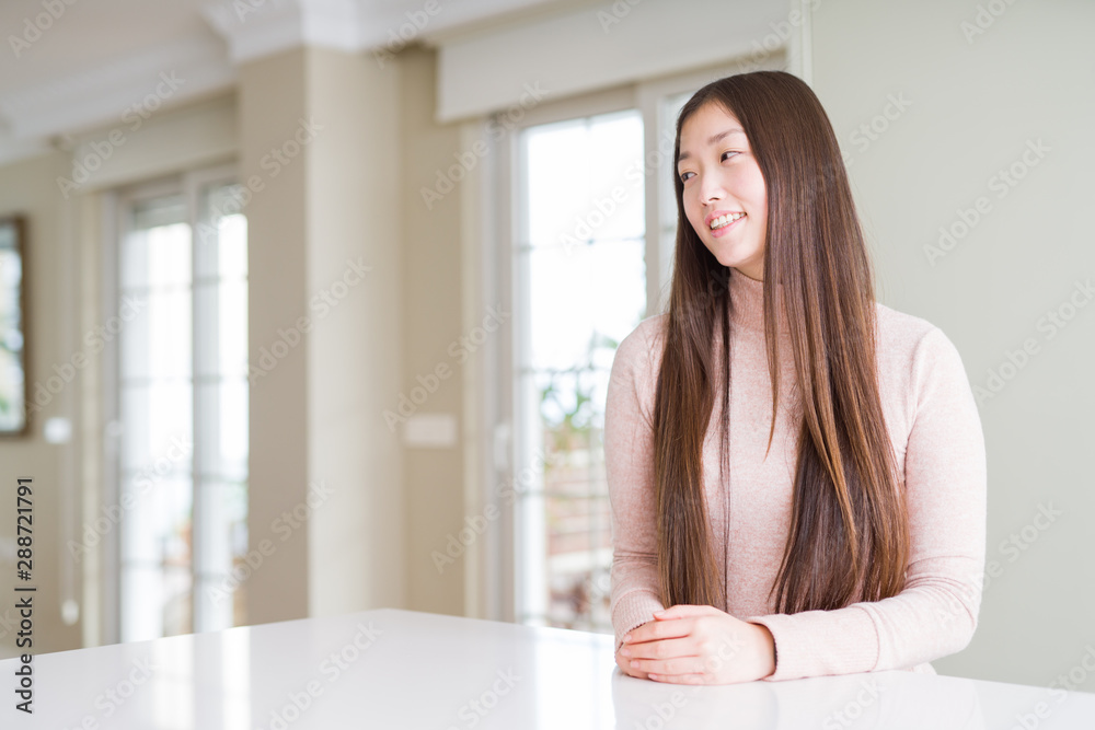 Beautiful Asian woman wearing casual sweater on white table looking away to side with smile on face, natural expression. Laughing confident. Beautiful Asian woman wearing casual sweater on white table looking away to side with smile on face, natural expression. Laughing confident.