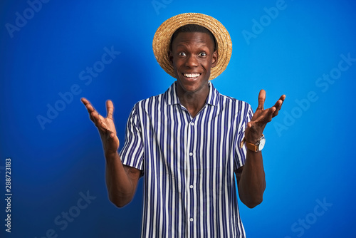 African american man wearing striped shirt and summer hat over isolated blue background celebrating crazy and amazed for success with arms raised and open eyes screaming excited. Winner concept