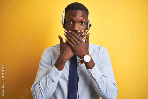 African american operator man working using headset over isolated yellow background shocked covering mouth with hands for mistake. Secret concept.