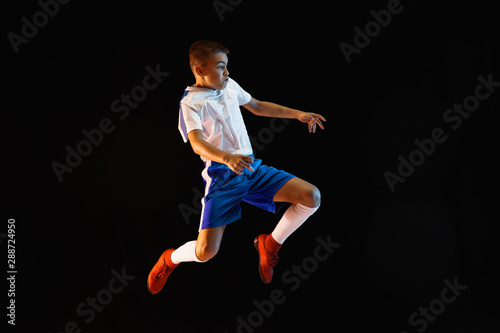 Young boy as a soccer or football player in sportwear making a feint or a kick with the ball for a goal on dark studio background. Fit playing boy in action, movement, motion at game. Purple neon