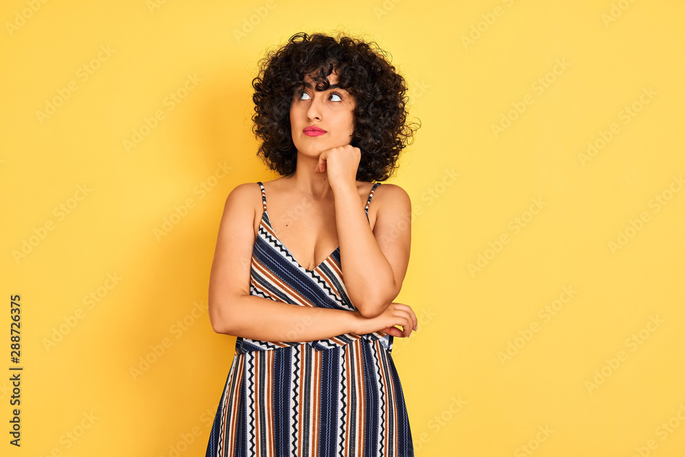 Arab woman with curly hair wearing striped colorful dress over isolated yellow background with hand on chin thinking about question, pensive expression. Smiling with thoughtful face. Doubt concept.