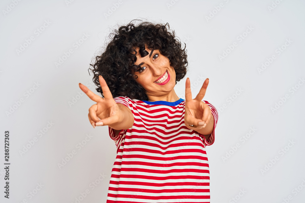 Young arab woman with curly hair wearing striped t-shirt over isolated white background smiling looking to the camera showing fingers doing victory sign. Number two. Young arab woman with curly hair wearing striped t-shirt over isolated white background smiling looking to the camera showing fingers doing victory sign. Number two.