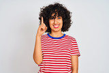 Young arab woman with curly hair wearing striped t-shirt over isolated white background showing and pointing up with finger number one while smiling confident and happy.