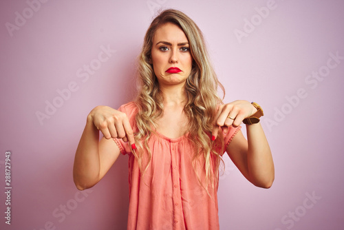 Young beautiful woman wearing t-shirt standing over pink isolated background Pointing down looking sad and upset, indicating direction with fingers, unhappy and depressed.