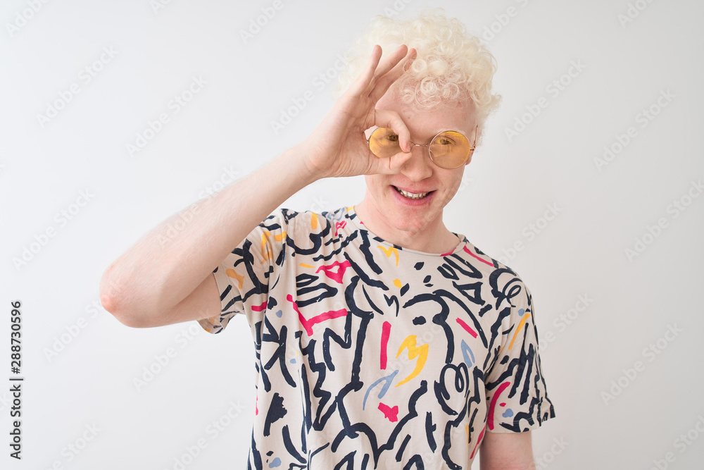 Young albino blond man wearing colorful t-shirt and sunglasses over isolated red background doing ok gesture with hand smiling, eye looking through fingers with happy face. Young albino blond man wearing colorful t-shirt and sunglasses over isolated red background doing ok gesture with hand smiling, eye looking through fingers with happy face.