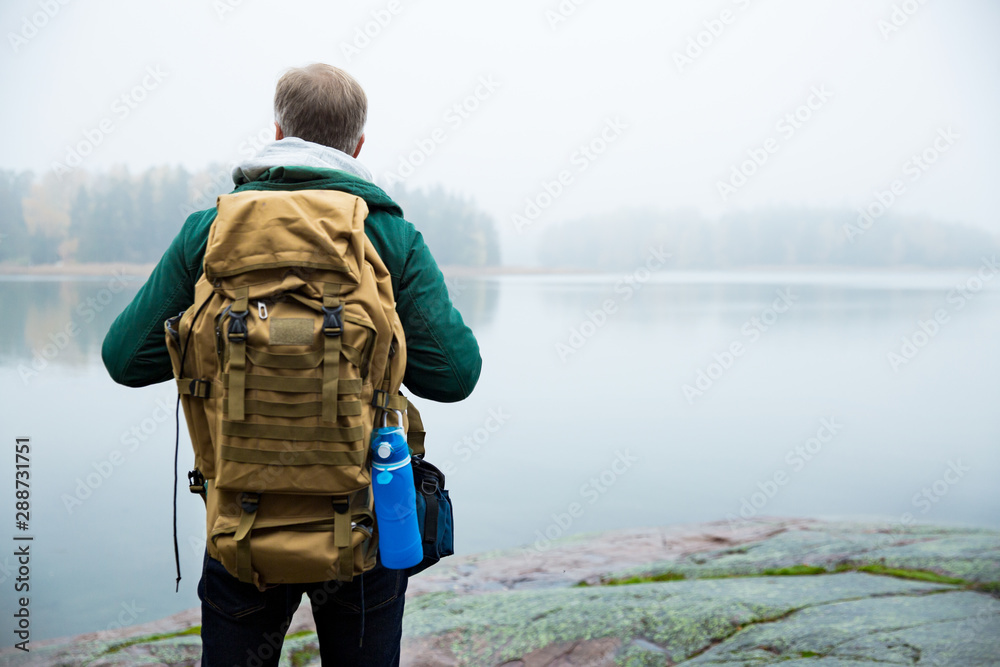 Mature man exploring Finland in the fall, looking into fog. Hiker with big backpack standing on mossy rock. Scandinavian landscape with misty sea and autumn forest. Back view - obrazy, fototapety, plakaty 
