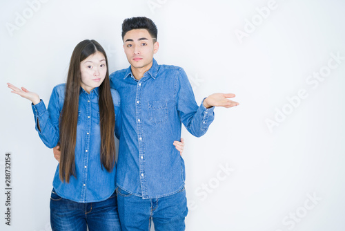 Beautiful young asian couple over white isolated background clueless and confused expression with arms and hands raised. Doubt concept.