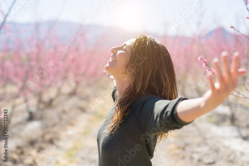 Beautiful middle age woman in the middle of pink peach flowers and trees smiling cheerful with open arms enjoying sunbathe on sunny day