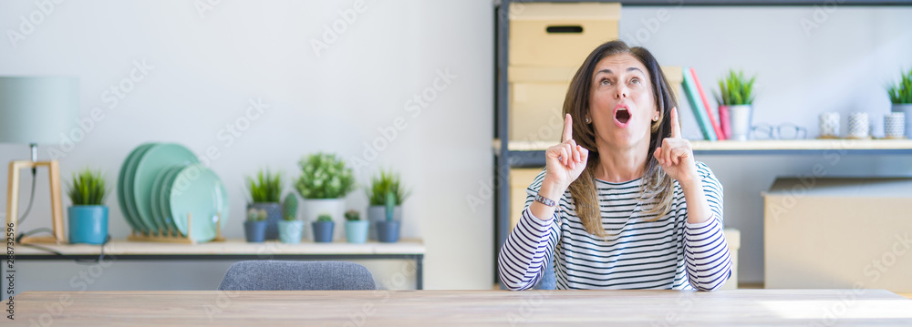 Wide angle photo of middle age senior woman sitting at the table at home amazed and surprised looking up and pointing with fingers and raised arms. Wide angle photo of middle age senior woman sitting at the table at home amazed and surprised looking up and pointing with fingers and raised arms.