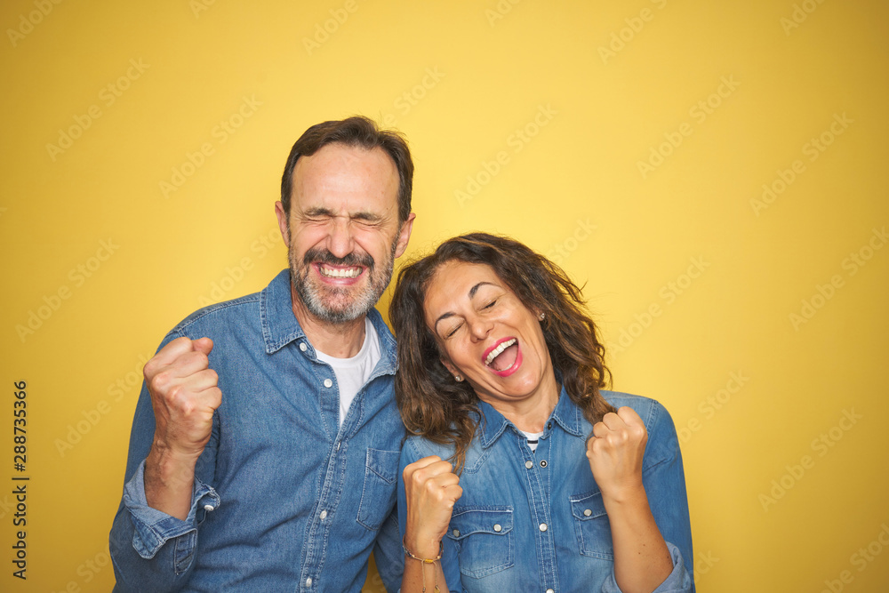 Beautiful middle age couple together standing over isolated yellow background very happy and excited doing winner gesture with arms raised, smiling and screaming for success. Celebration concept. Beautiful middle age couple together standing over isolated yellow background very happy and excited doing winner gesture with arms raised, smiling and screaming for success. Celebration concept.