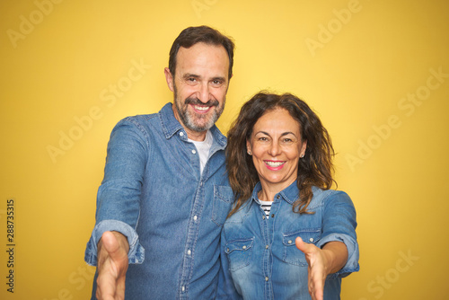 Beautiful middle age couple together standing over isolated yellow background smiling friendly offering handshake as greeting and welcoming. Successful business.