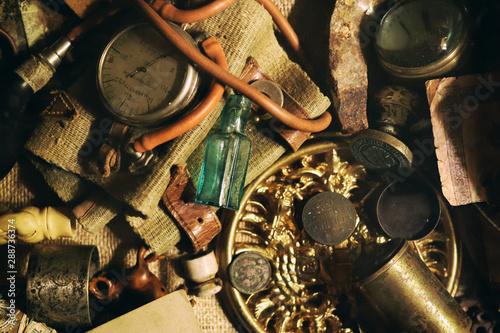 Different antique items on the table: bronze jewelry, old money, retro manometer, magnifier, glass bottle, silverware. Vintage background from a collection of antiques. Close-up selected focus