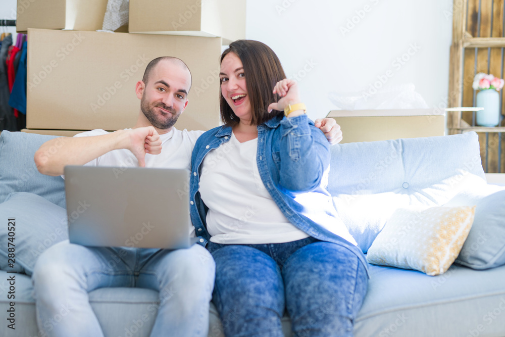 Young couple sitting on the sofa arround cardboard boxes moving to a new house using laptop with surprise face pointing finger to himself Young couple sitting on the sofa arround cardboard boxes moving to a new house using laptop with surprise face pointing finger to himself