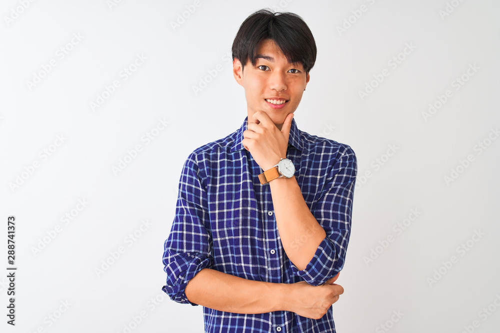 Young chinese man wearing casual blue shirt standing over isolated white background looking confident at the camera with smile with crossed arms and hand raised on chin. Thinking positive. Young chinese man wearing casual blue shirt standing over isolated white background looking confident at the camera with smile with crossed arms and hand raised on chin. Thinking positive.