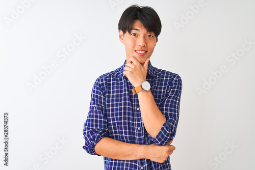 Young chinese man wearing casual blue shirt standing over isolated white background looking confident at the camera with smile with crossed arms and hand raised on chin. Thinking positive.