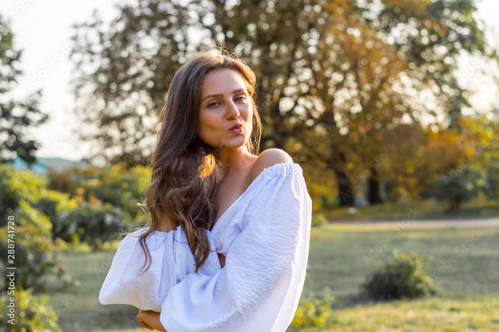Young beautiful woman with bare shoulders in white clothes blows a kiss in the park at the sunset Young beautiful woman with bare shoulders in white clothes blows a kiss in the park at the sunset