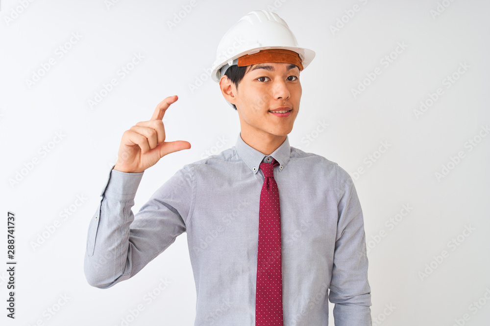 Chinese architect man wearing tie and helmet standing over isolated white background smiling and confident gesturing with hand doing small size sign with fingers looking and the camera. 