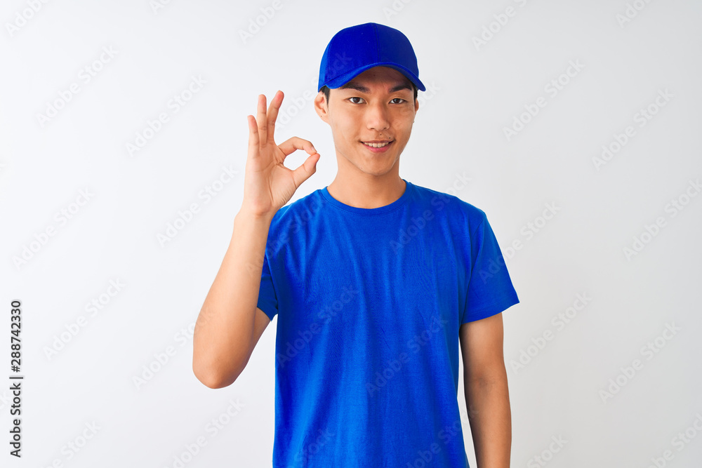 Chinese deliveryman wearing blue t-shirt and cap standing over isolated white background smiling positive doing ok sign with hand and fingers. Successful expression.